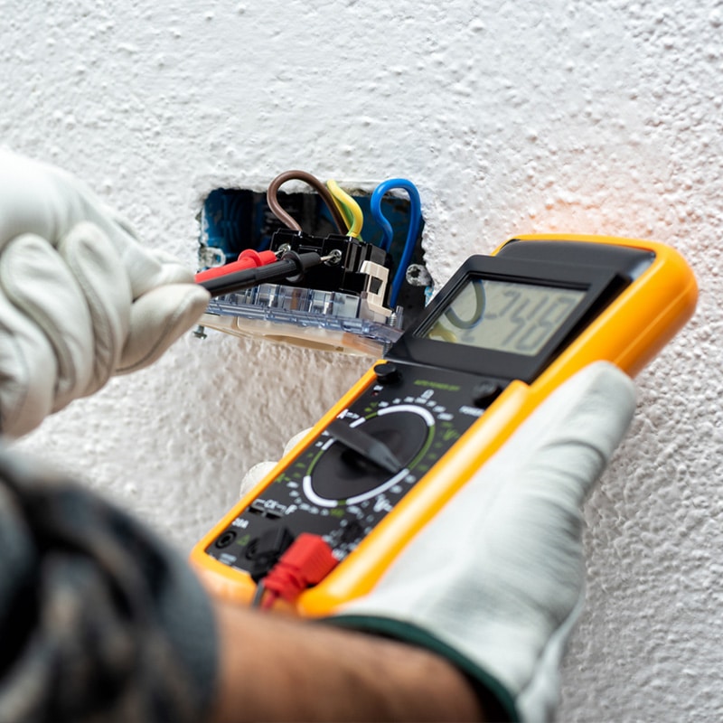 Electrician using a multimeter to measure voltage of wall socket wiring for safety check.