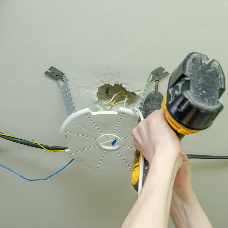 Installing a ceiling light fixture with a power drill, wires exposed for electrical setup.