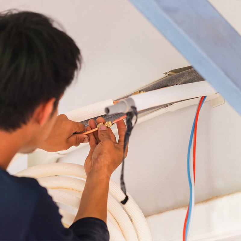 Technician installing air conditioning unit with copper pipes and insulation materials indoors.