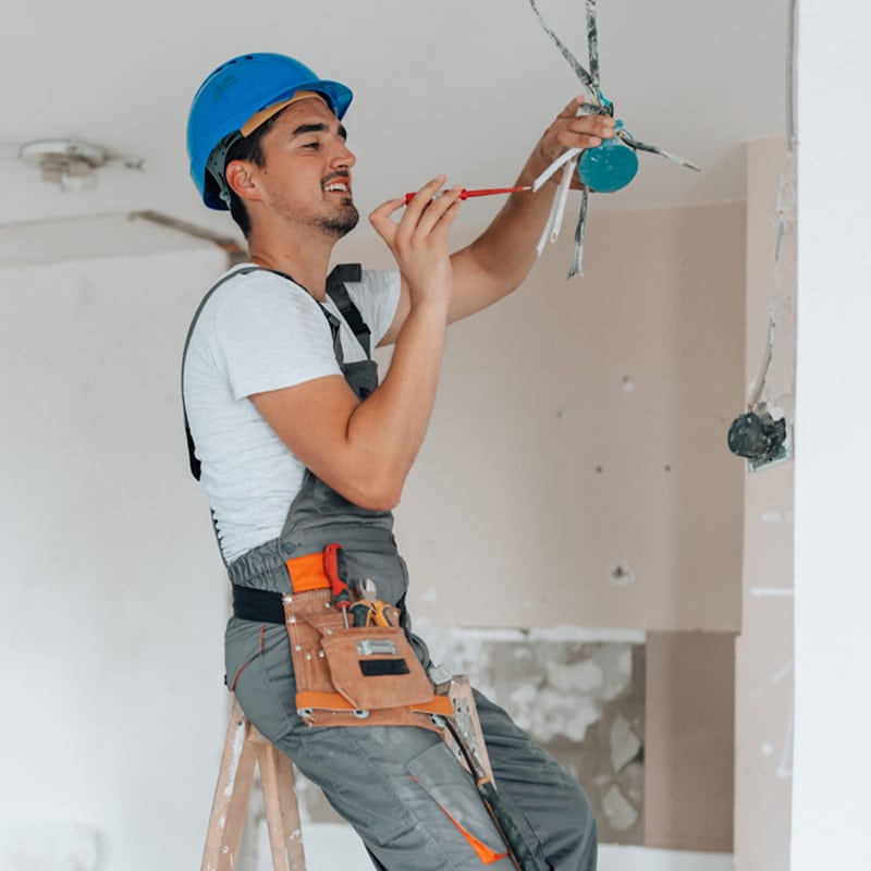 Electrician on ladder fixing ceiling wires, wearing blue helmet and tool belt in under-construction room.
