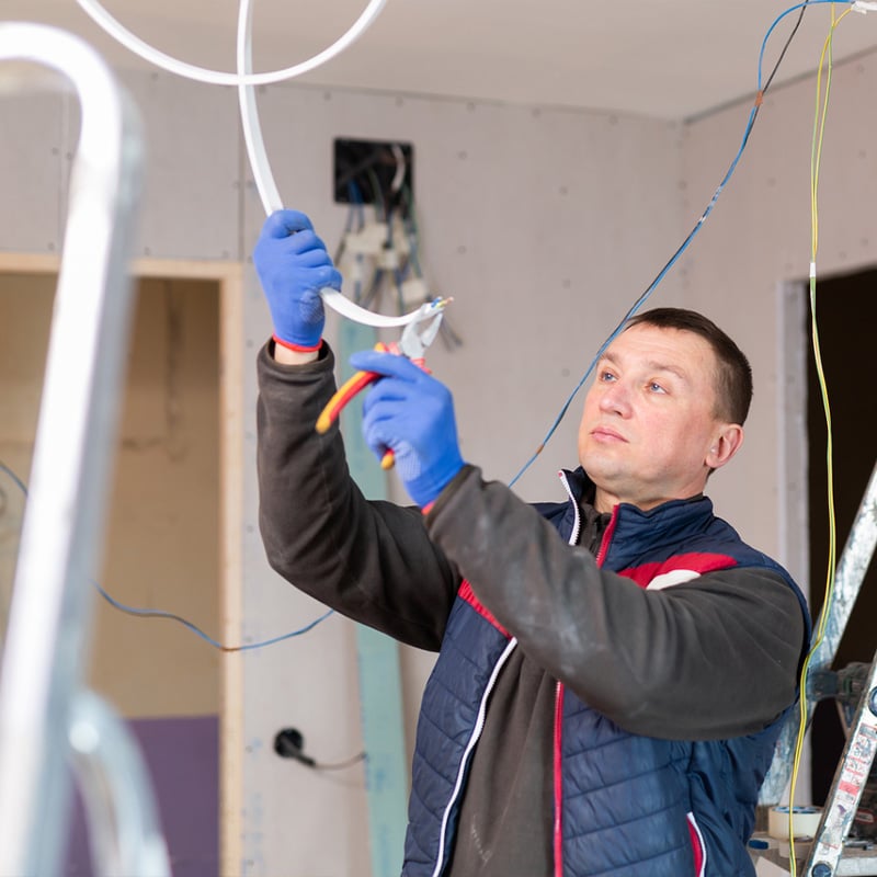 Electrician using tools to install wiring in a home under construction, wearing gloves and protective gear.