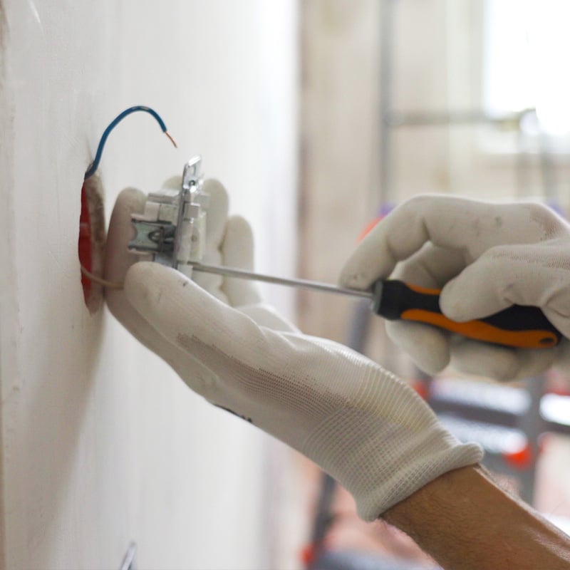 Electrician installing wall outlet with screwdriver and gloves.