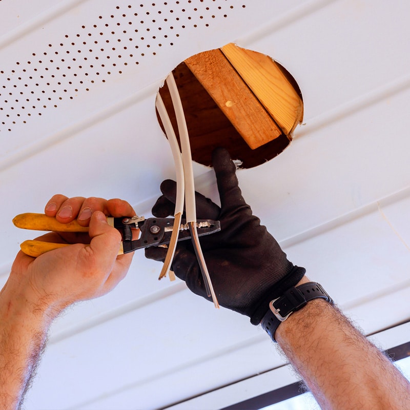 Electrician stripping wires through a ceiling hole with wire strippers and gloved hand, preparing for installation.
