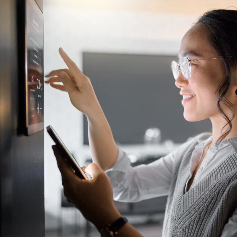 Woman adjusting smart home thermostat with a smartphone, smiling and interacting with touchscreen.