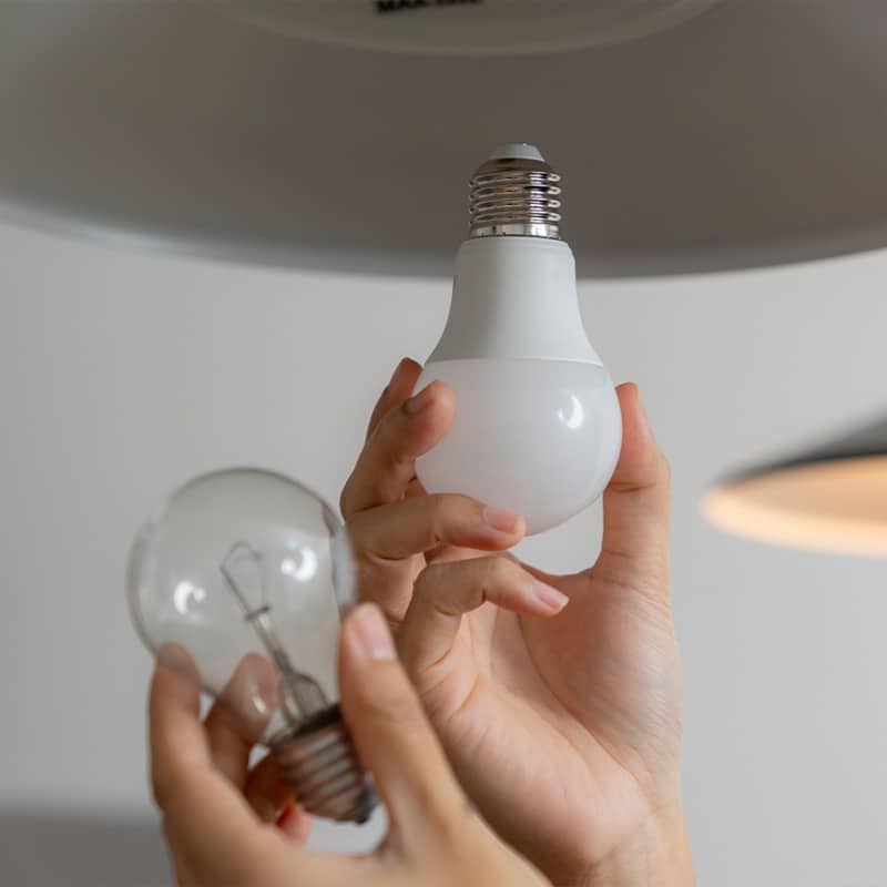 Hand replacing incandescent bulb with energy-efficient LED light in a lamp fitting, promoting sustainability.