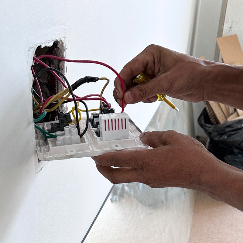 Electrician fixing a wall socket with red and yellow wires using a screwdriver.