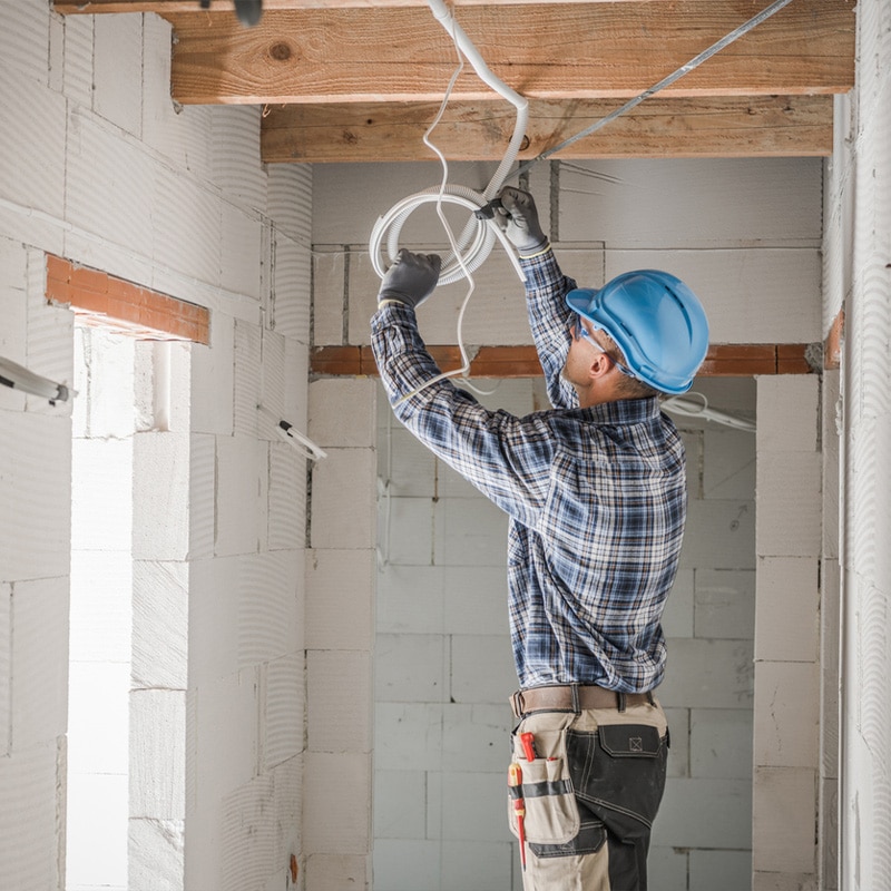 Electrician installing wiring in a construction site, wearing a blue hard hat and plaid shirt.