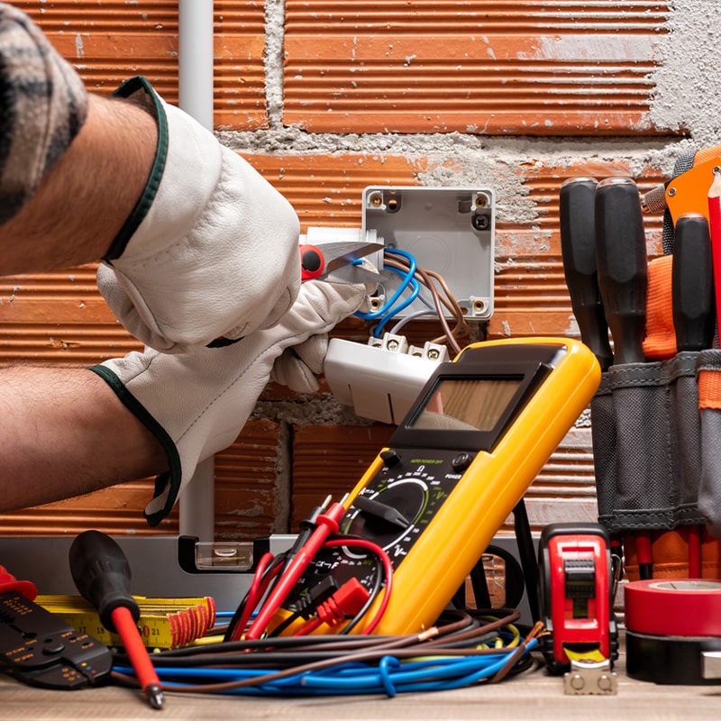 Electrician repairing outlet, using tools and multimeter, with a toolbox on brick wall background.