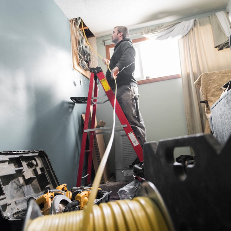 Electrician on ladder installing wiring in a room, tools on the floor around him.