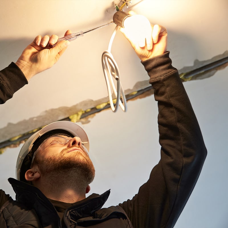Electrician wearing a hard hat installs a ceiling light, using a screwdriver for wiring.
