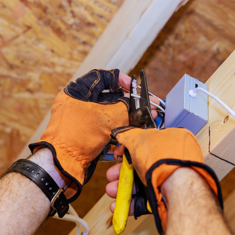 Electrician wearing gloves installs electrical wiring in a wooden frame with pliers.