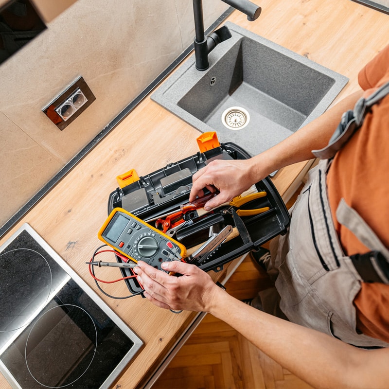 Handyman using a multimeter from a toolbox on a kitchen countertop near a sink and stovetop.
