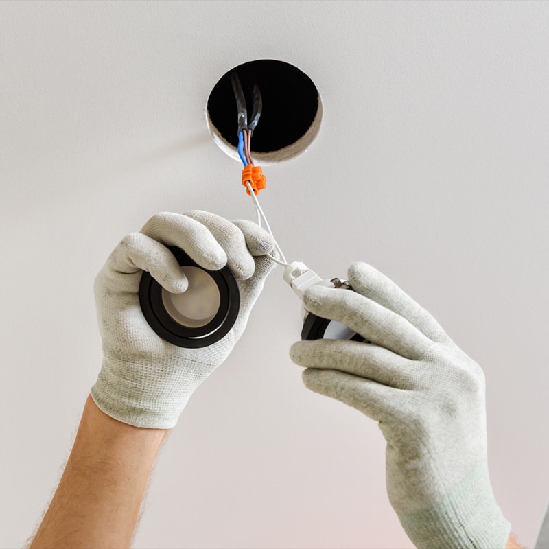 Electrician installing a recessed ceiling light fixture, wearing safety gloves, connecting wires for home lighting.