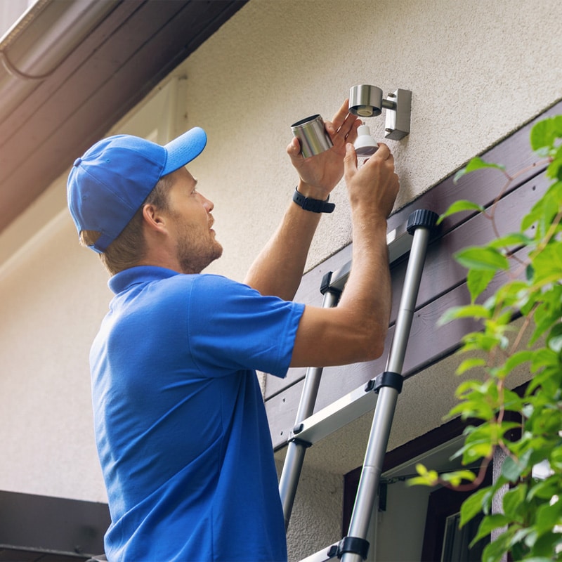Technician installs outdoor security camera on house wall using ladder, ensuring home safety and surveillance.